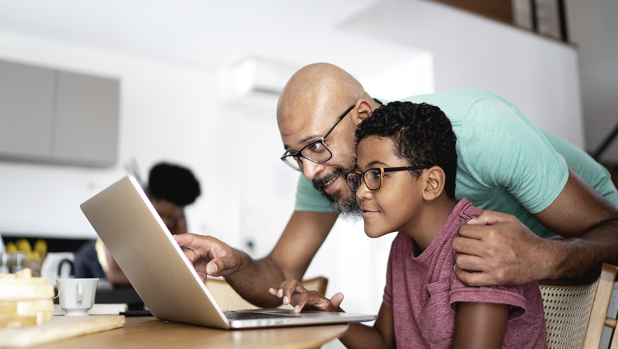 A picture of a boy using a laptop and his father is looking over his shoulder.